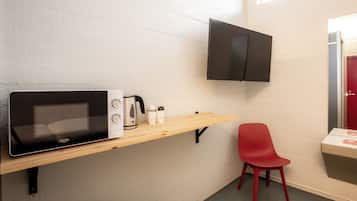 A kitchenette with a microwave, kettle, and coffee maker on a wooden shelf, a red chair, and a flat-screen TV mounted on the wall.