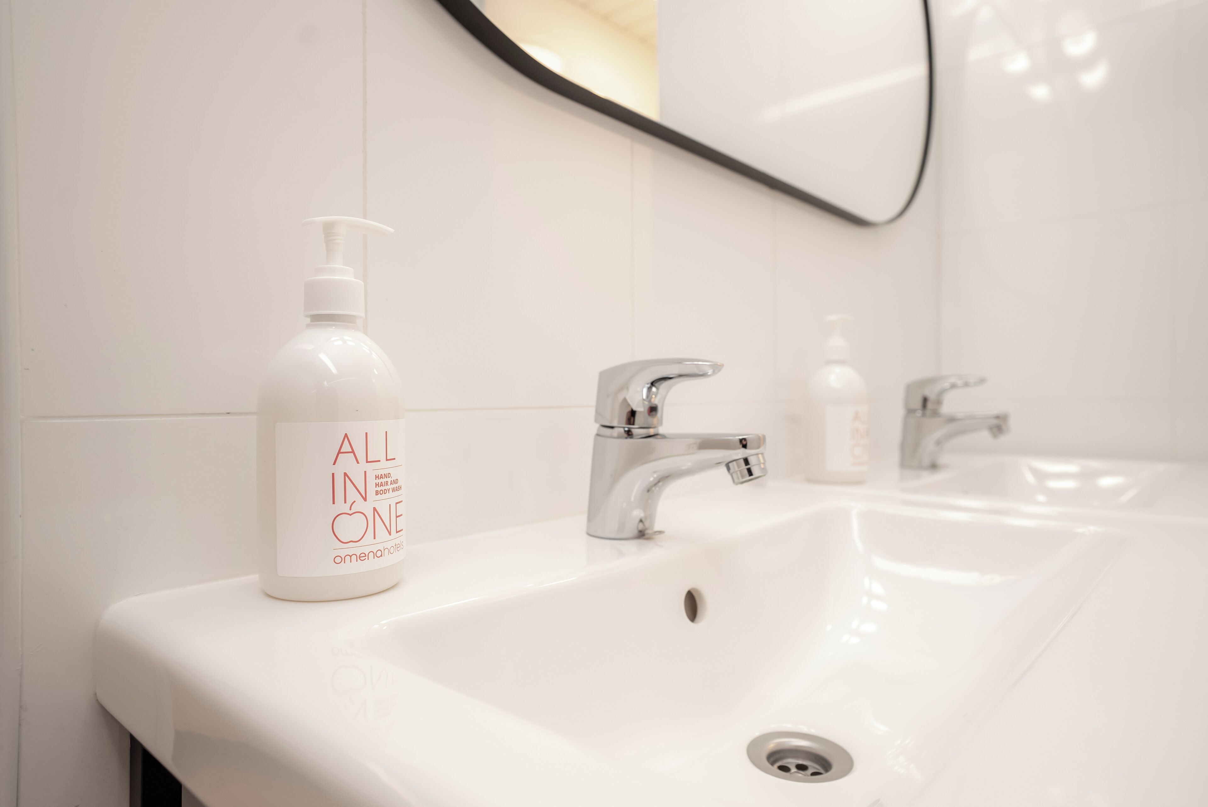 A bathroom with a white sink, two faucets, and a bottle of 'ALL IN ONE' hand soap.