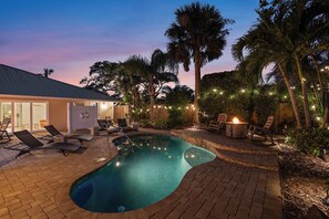 A swimming pool with lounge chairs and a fire pit, surrounded by palm trees and string lights.