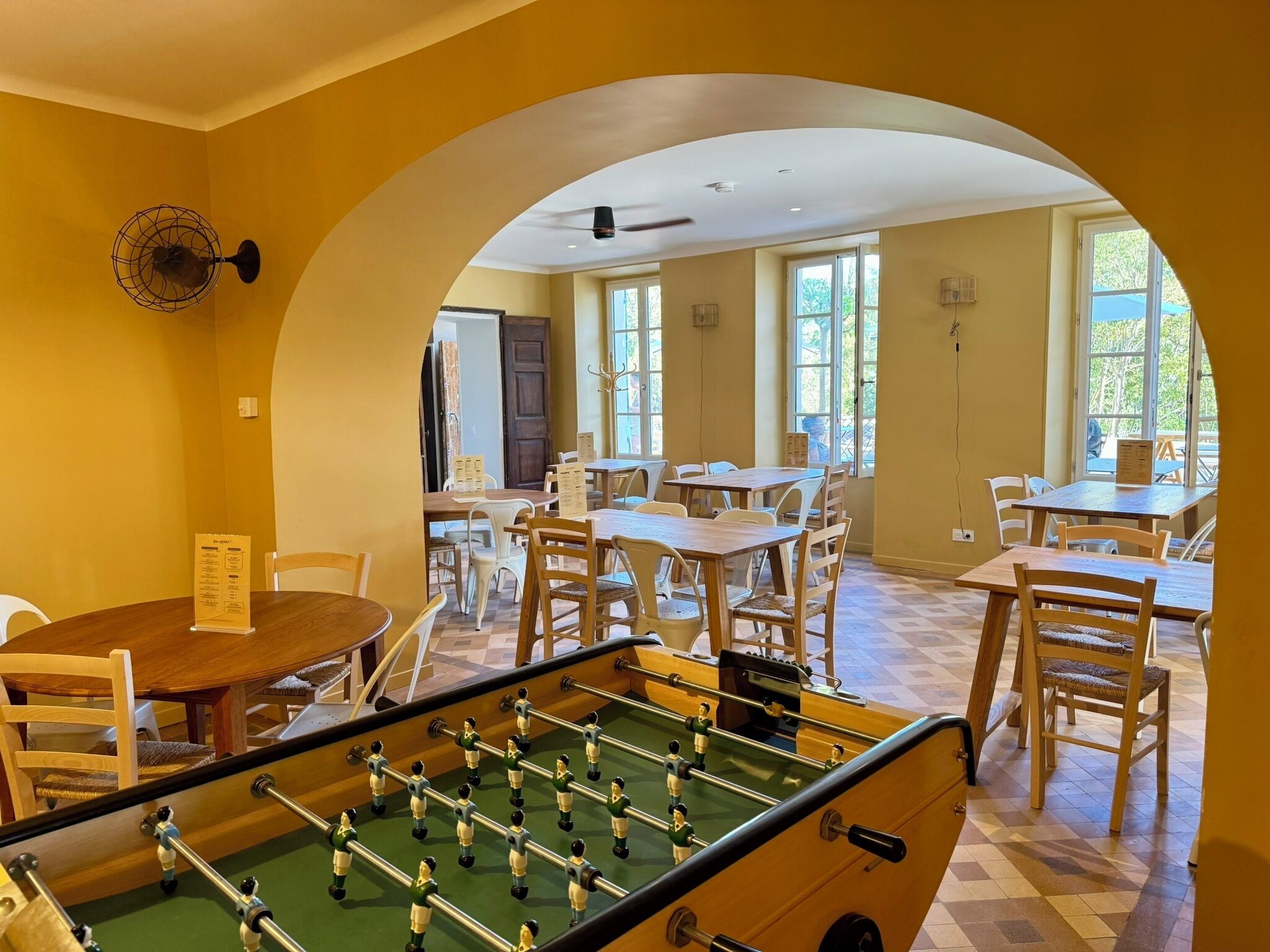 A room with a foosball table, multiple tables and chairs, and large windows allowing natural light.