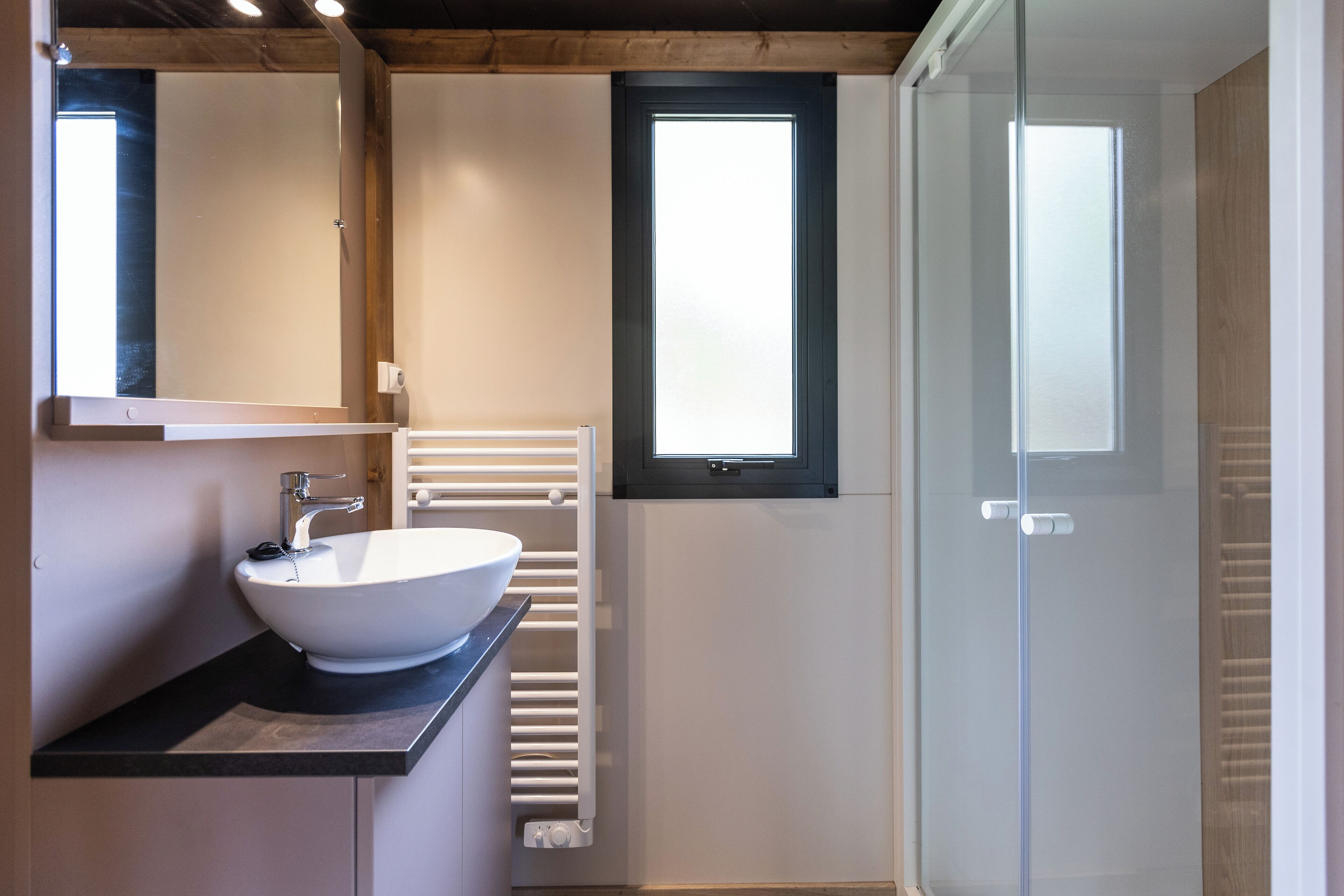 A modern bathroom with a white sink, a mirror, a window, and a glass shower enclosure.