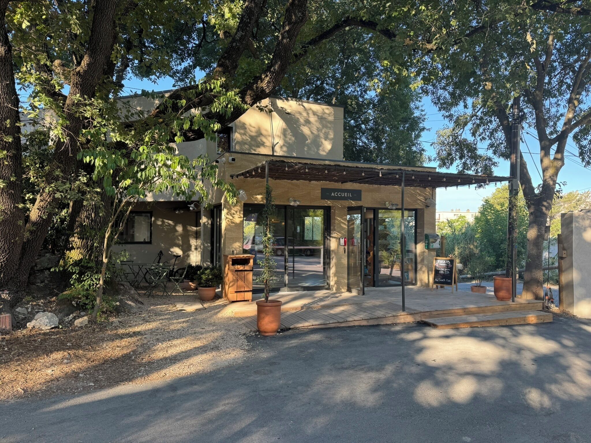 A building with a covered entrance labeled "ACCUEIL," surrounded by trees and potted plants.