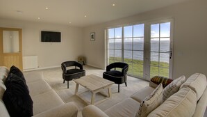 Ein großzügiges Wohnzimmer mit einer großen Couch, zwei Sesseln, einem Couchtisch und Meerblick durch große Glasfenster.