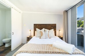 A bedroom with a large bed, a wooden headboard, two bedside tables with lamps, and a view of the outdoors through a sliding door. - Studio Apartment in Colourful Bo-kaap (Cape Town)