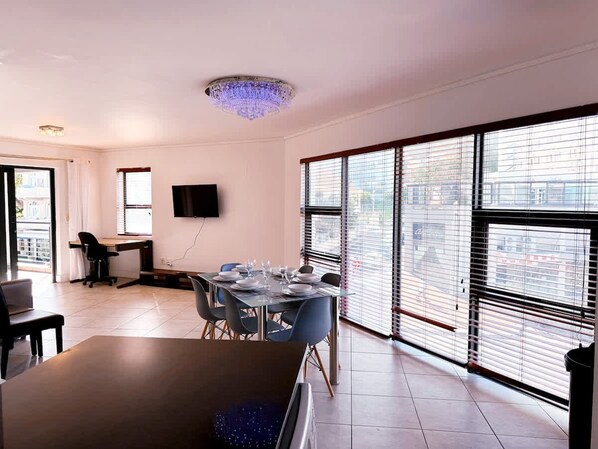 A modern dining area with a table set for six, a TV mounted on the wall, and large windows with blinds. - Spacious 1 Bedroom Apartment Atlantica Seapoint (Cape Town)