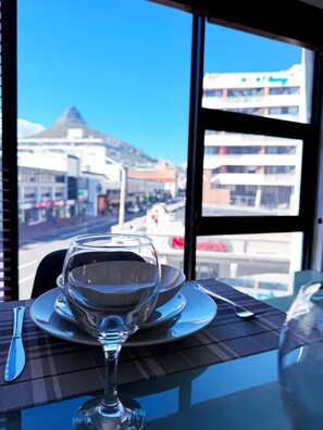 A table with a wine glass, a plate, and cutlery, set against a window with a view of buildings and a mountain. - Spacious 1 Bedroom Apartment Atlantica Seapoint (Cape Town)