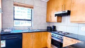 A compact kitchen with wooden cabinets, a gas stove, a dishwasher, and a window providing natural light. - Spacious 1 Bedroom Apartment Atlantica Seapoint (Cape Town)