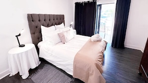 A neatly made bed with a tufted headboard, a bedside table with a lamp, and a view of the outdoors through a window with dark curtains. - Spacious 1 Bedroom Apartment Atlantica Seapoint (Cape Town)