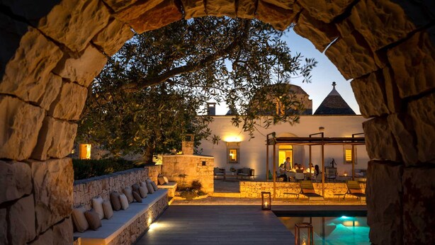 A stone archway leading to a pool area with a lounge chair and a stone bench.