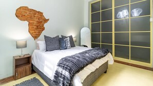 A bedroom with a brick wall art piece, a bed with a white and gray striped blanket, a nightstand with a lamp, and a large window with frosted glass panels.
