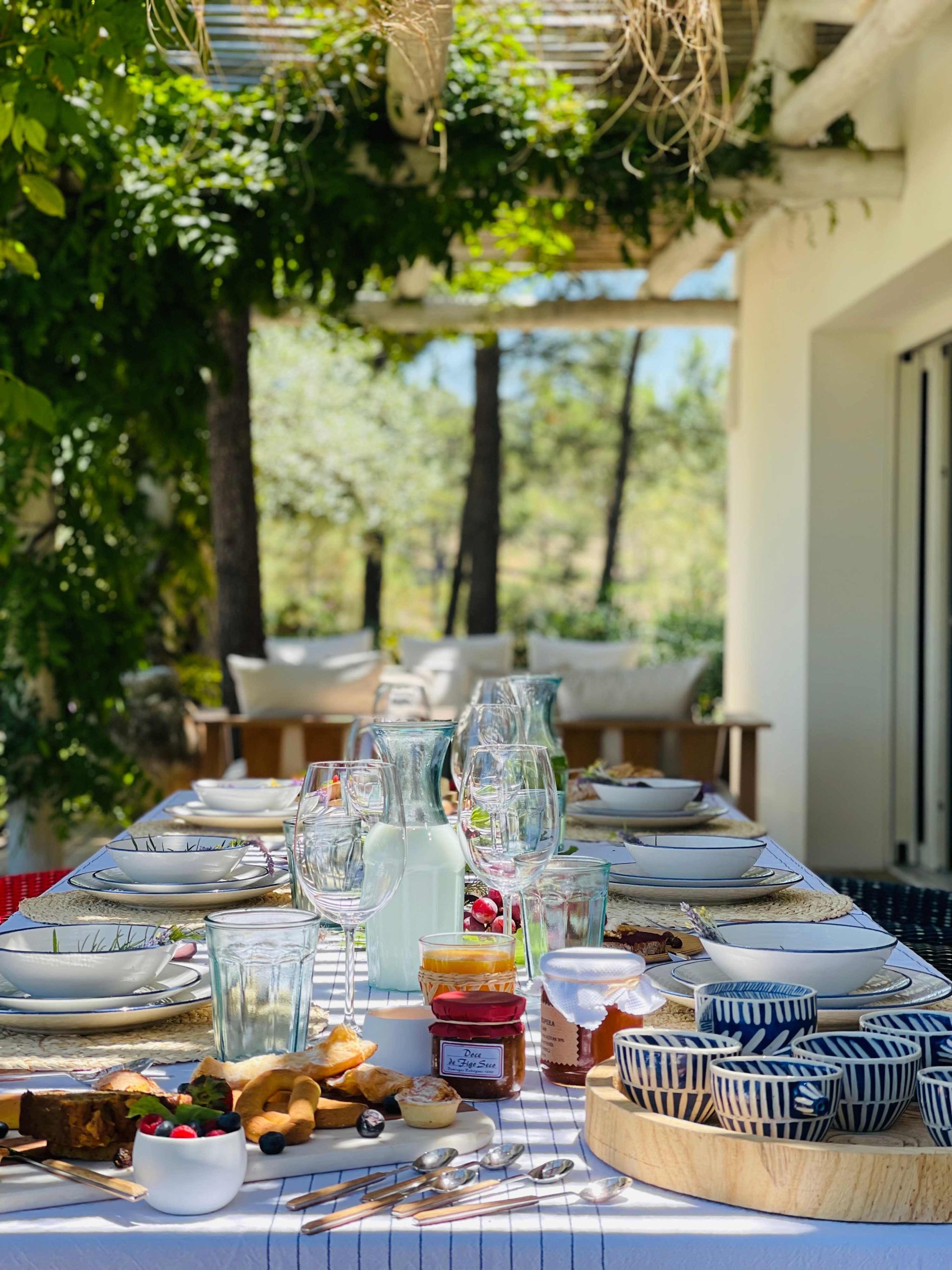 An outdoor dining table set with plates, glasses, and food items, surrounded by greenery and trees.