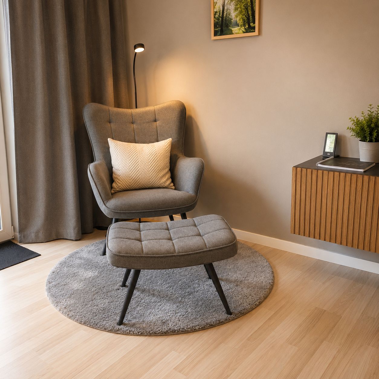 Ein modernes Zimmer mit einem grau gepolsterten Sessel, passendem Hocker, einer Stehlampe, einem kleinen Holznachttisch mit Pflanze und einem gerahmten Bild an der Wand.