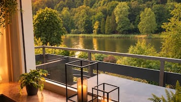 Ein Balkon mit Blick auf einen See und einen Wald, ein Tisch mit Kerzen und ein Topf mit einer Pflanze.