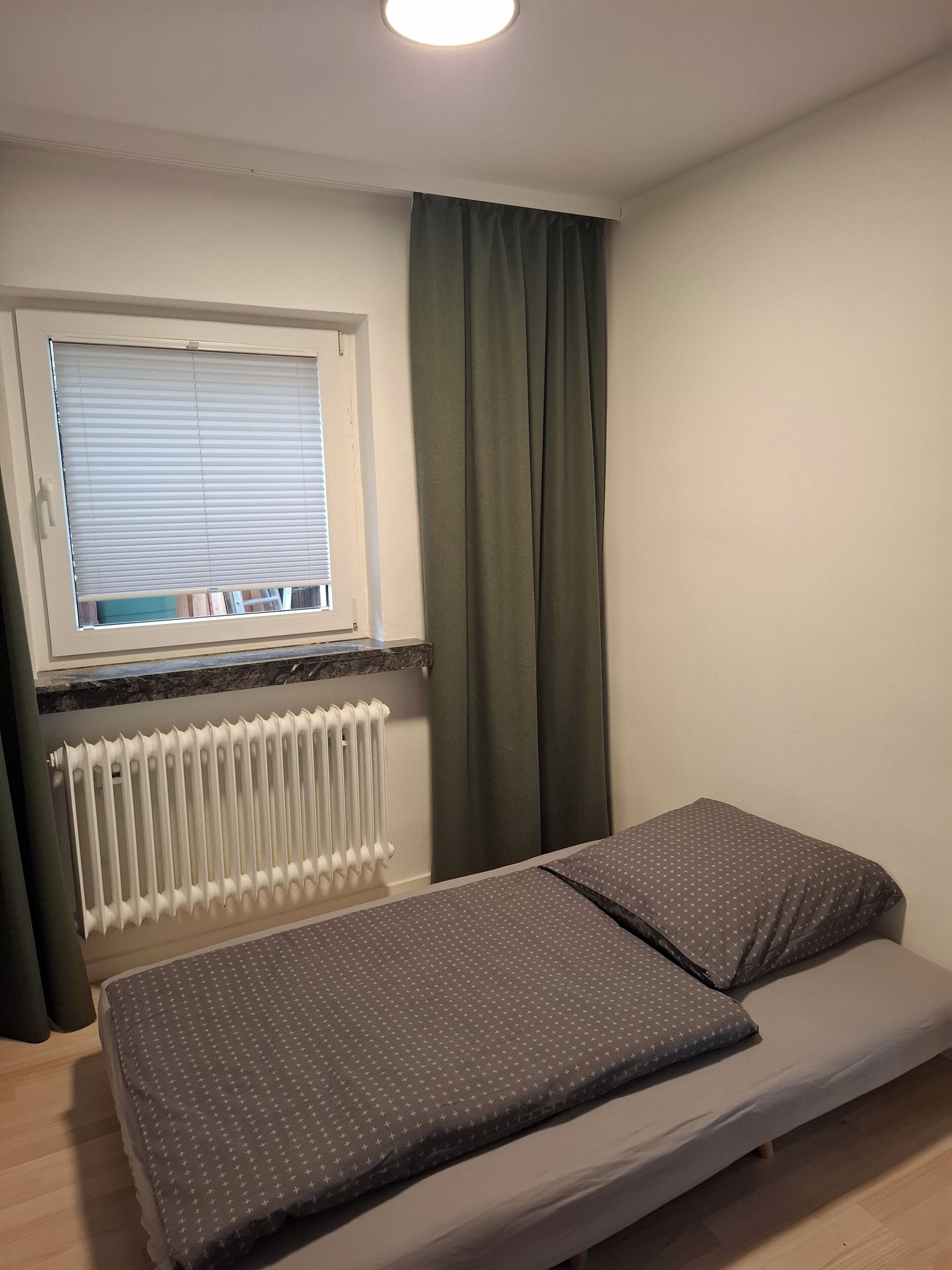 Une petite pièce comprenant un lit, une fenêtre avec des stores, un radiateur et un rideau.