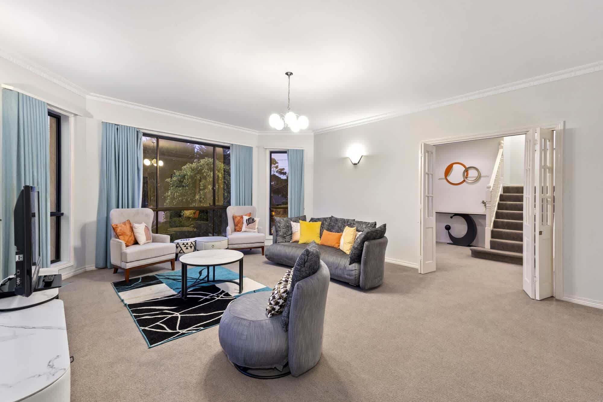 Ein modernes Wohnzimmer mit zwei Sofas, einem Couchtisch und einem Fernseher. Ein großes Fenster mit Vorhängen, eine Treppe und eine Wand mit abstrakter Kunst sind ebenfalls zu sehen.
