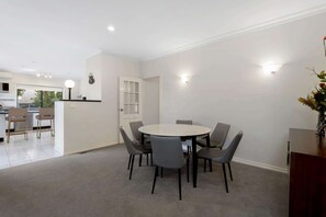 A dining area with a round table and six chairs, a kitchen in the background, and a door leading to another room. - Fairhaven (Mount Martha)