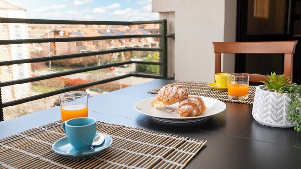 Un balcón con una mesa dispuesta para el desayuno, con cruasanes, un vaso de zumo de naranja y una taza.