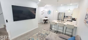 Una sala moderna con televisor de pantalla plana, un comedor con mesa y sillas, una cocina con mostrador tipo bar, y una alfombra estampada en el suelo.