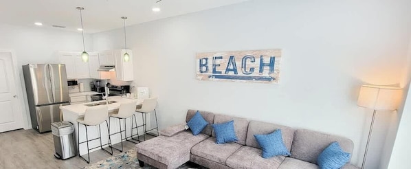 Una cucina moderna con elettrodomestici in acciaio inossidabile, un'area pranzo con sgabelli da bar, un divano grigio con cuscini blu e un cartello con la scritta 'BEACH'.