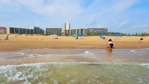 Una playa con edificios al fondo, una orilla arenosa y olas acercándose a la costa.