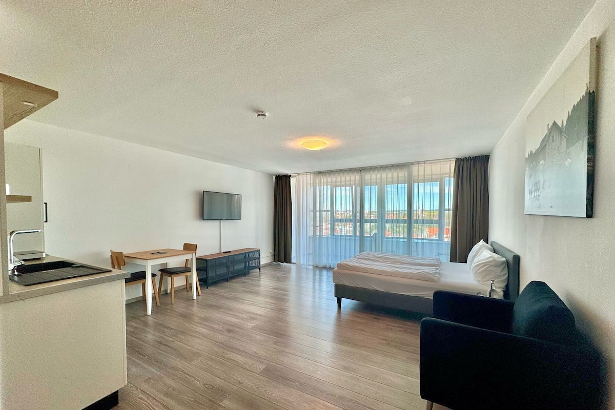 A hotel room with a bed, a desk, a chair, a TV, and a view of the city.