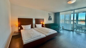 Hotellihuone, jossa on suuri sänky, kaksi yöpöytävalaisinta, vihreä nojatuoli, pieni pöytä ja parveke, jolta on näkymä.
