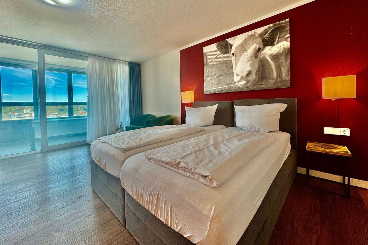 A hotel room with two beds, a large window with a view, a green chair, and a wall-mounted lamp.
