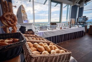 A buffet setup with various breads and pastries, a coffee machine, and a view of the city through large windows. - My Park hotel Kempten by AR hotels (Kempten)