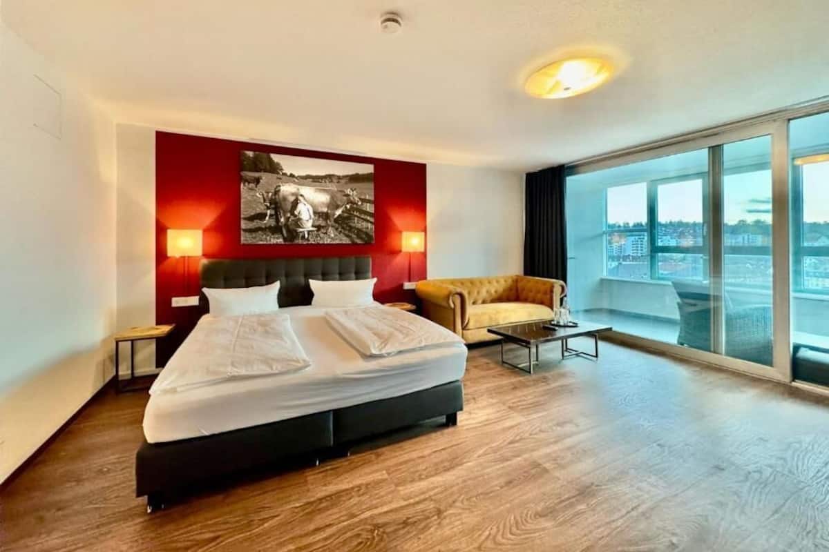 A modern hotel room with a large bed, two bedside lamps, a sofa, a coffee table, and a large window offering a city view.