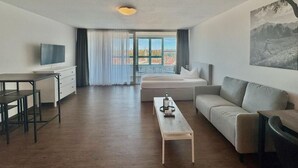 Moderni hotellihuone, jossa on sänky, sohva, televisio, työpöytä ja suuri ikkuna, josta on näkymä.