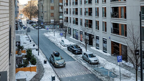 Une rue moderne en milieu urbain, recouverte de neige, avec un grand immeuble dont on aperçoit le logo « UNIQA » en arrière-plan, et plusieurs voitures garées.