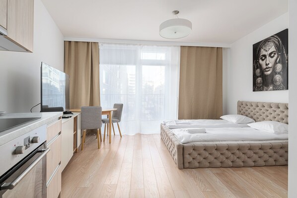 Una habitación de hotel moderna con kitchenette, una cama, zona de comedor y un ventanal con cortinas.