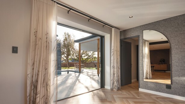 A spacious room with a large sliding glass door leading to an outdoor area, a herringbone-patterned wooden floor, and a kitchen visible through an archway.