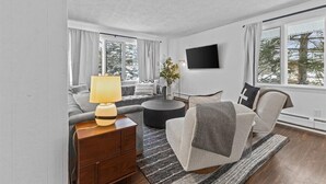 Een moderne woonkamer met een bank, fauteuil, rond bijzettafeltje en een aan de muur bevestigde televisie.