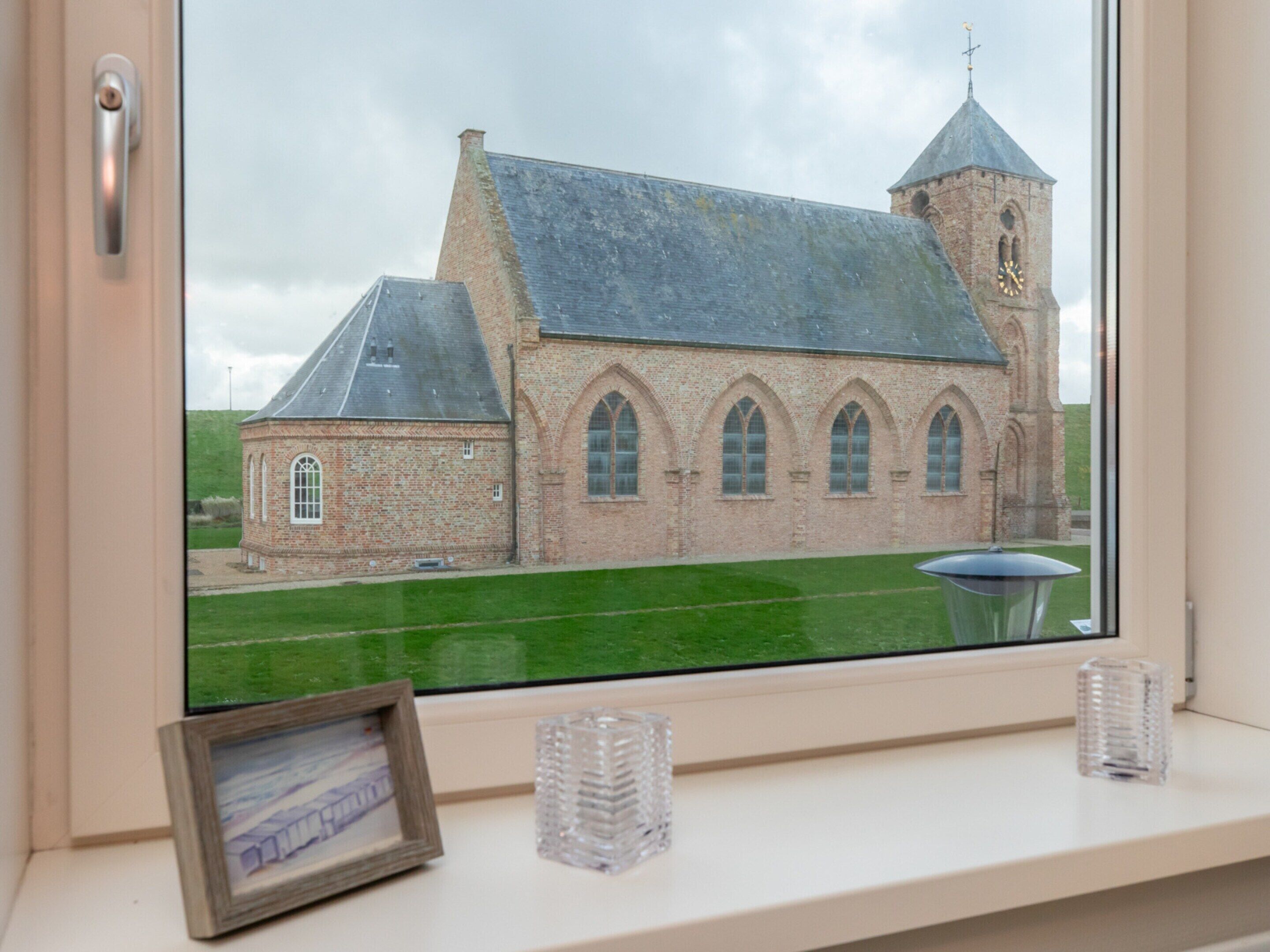 A view of a church with a clock tower and a smaller building, through a window with a picture frame and two glass candle holders.