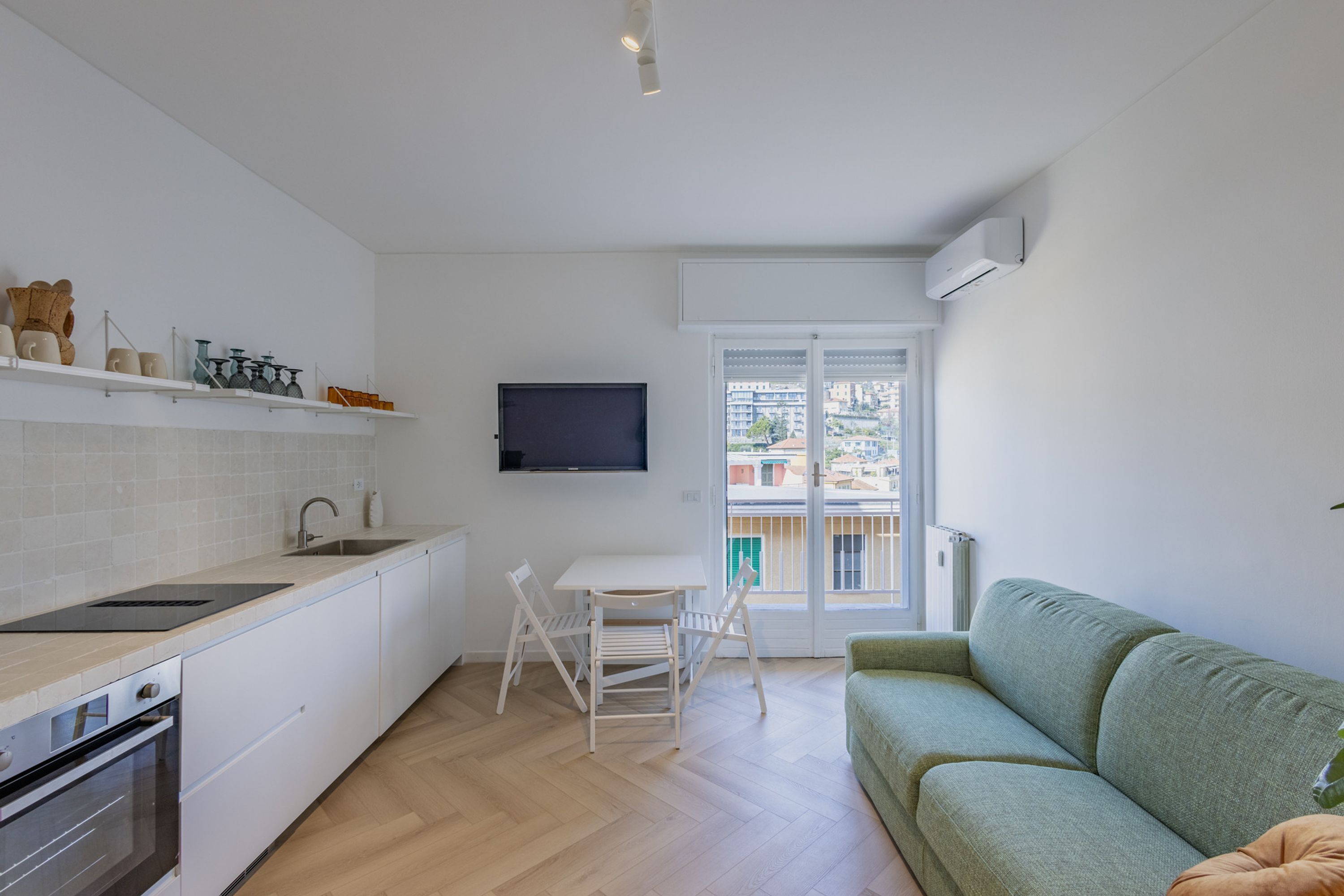 A modern kitchen with white cabinetry, a built-in oven, and a dining area with a white table and chairs. A green sofa is positioned near a sliding glass door leading to a balcony.