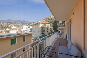 A balcony with a sofa and a table, overlooking a residential area with buildings and a mountain in the background. - Goethe Charming Home by Wonderful Italy (Sanremo)