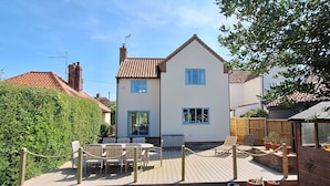 A two-story house with a white exterior and a tiled roof. It features a wooden deck with outdoor furniture and potted plants. - Tern Cottage (Holme-next-the-Sea), Norfolk (Holme-next-the-Sea)
