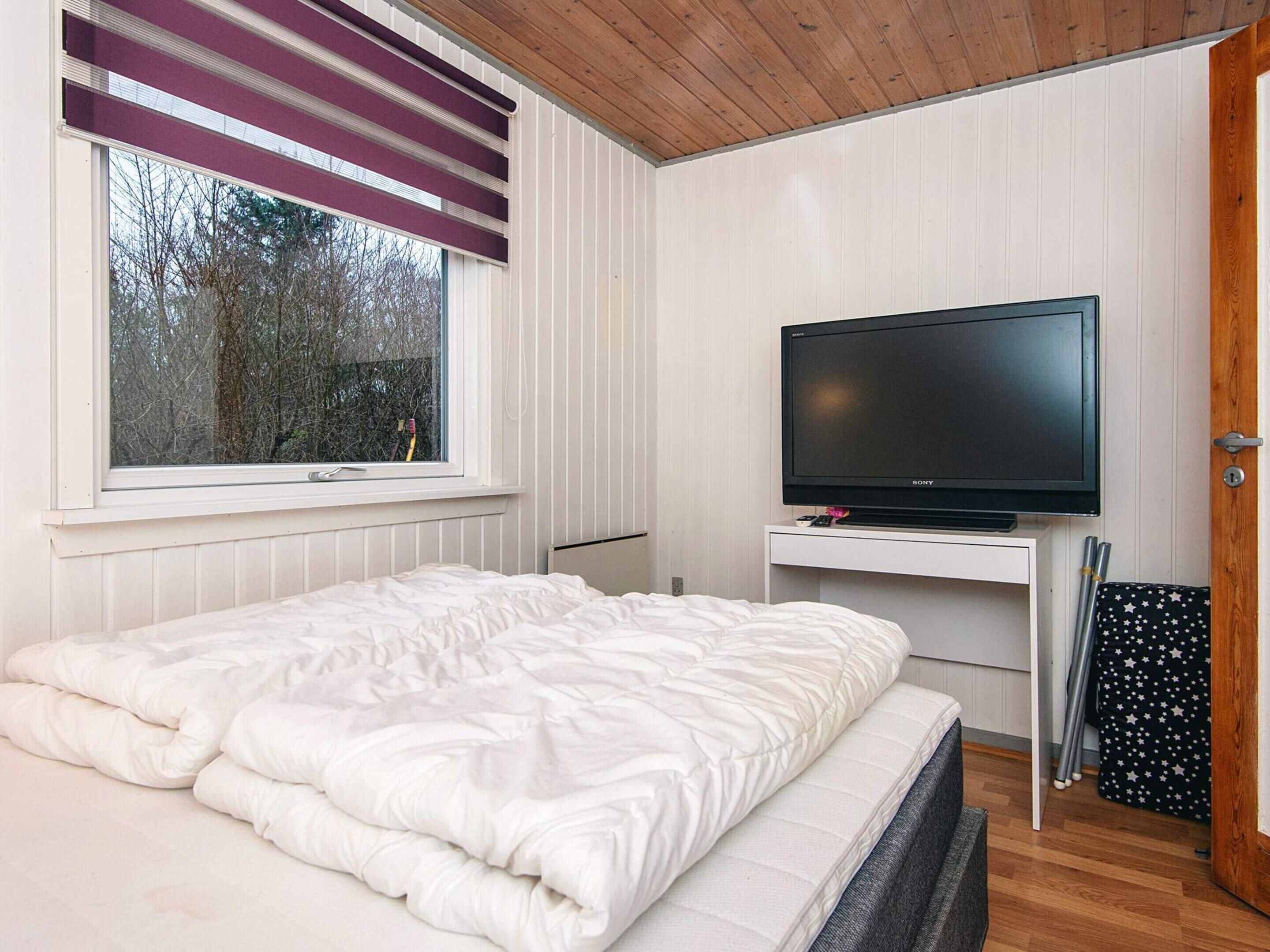 A bedroom with a bed, a television on a stand, a window with blinds, and a door.