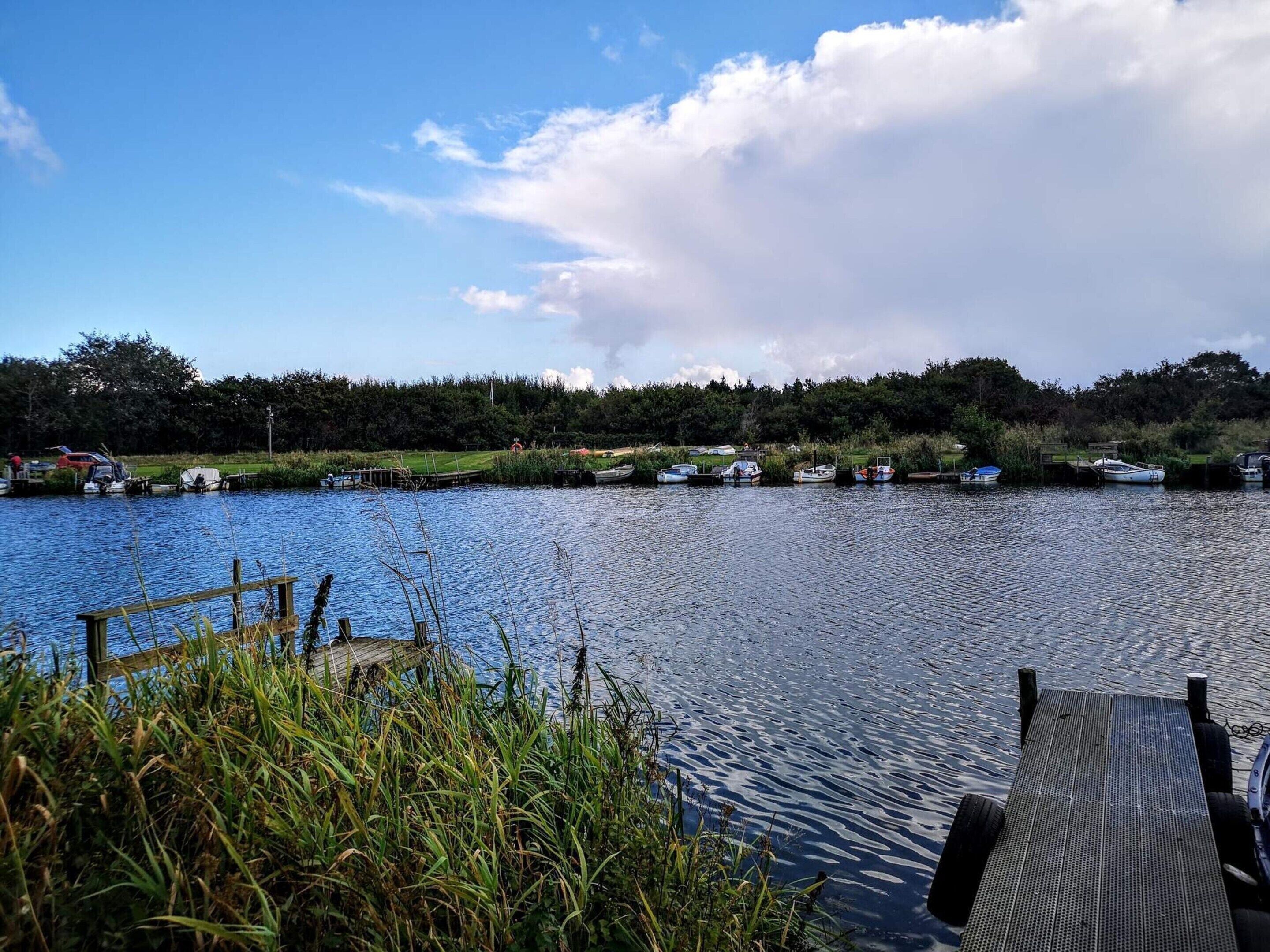 Un lago tranquillo con barche ormeggiate lungo la riva, circondato da vegetazione e un cielo sereno.