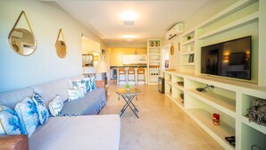 A modern living room with a sofa, coffee table, and TV. There is a kitchen in the background with a dining area. - Bz09 Quadra da praia Casa 5 suites (Buzios)