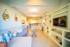 Una sala de estar moderna con sofá, mesa de centro y televisor. Se ve una cocina al fondo con zona de comedor.