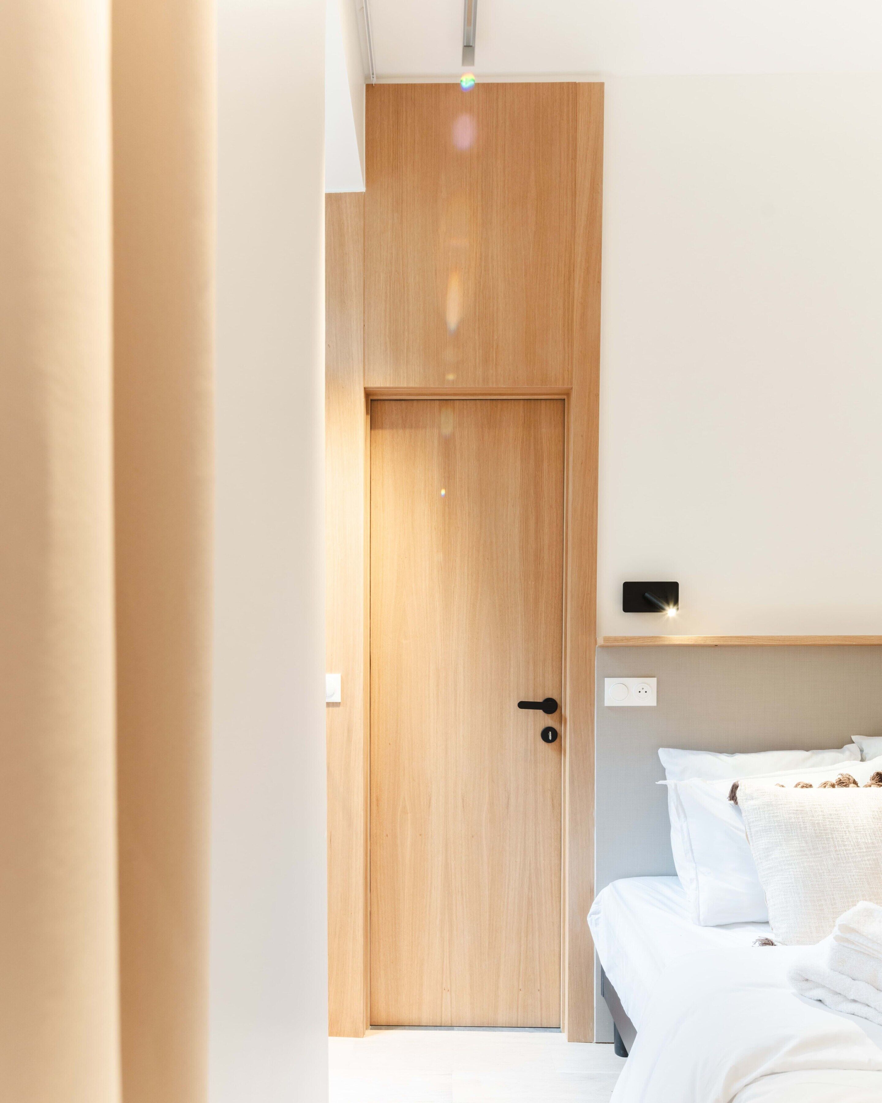 Een hotelkamer met een houten deur, een bed met witte beddengoed en een klein plankje boven het bed.
