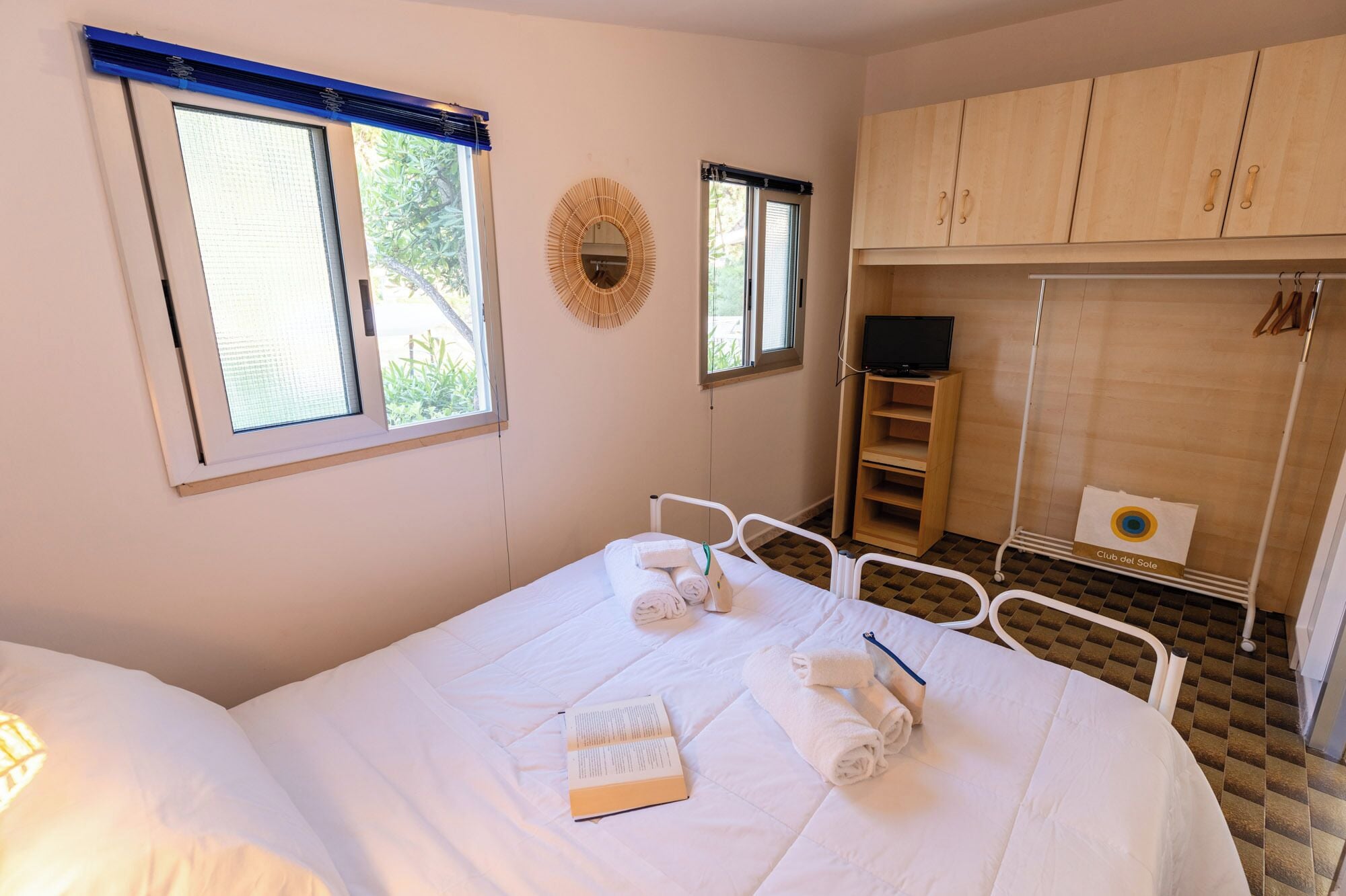 Habitación de hotel con cama, dos ventanas, televisor, armario y un pequeño estante con una toalla y un libro.