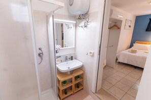 Une salle de bain avec une douche, un lavabo et un miroir. On y trouve une étagère en bois avec une plante et une bouteille. La pièce est équipée d’un lit recouvert d’une courtepointe blanche et d’un mur bleu.