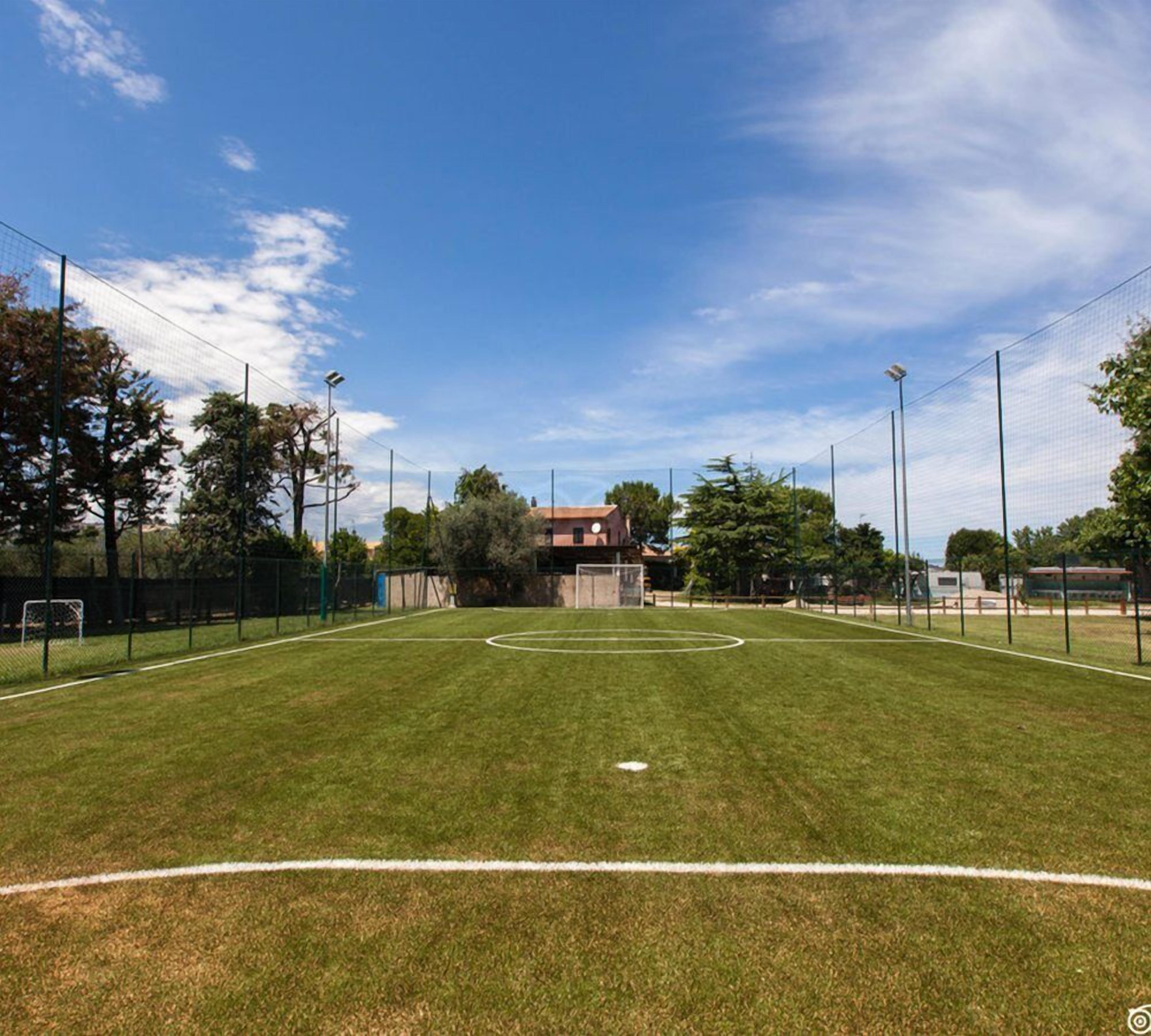 A soccer field with goalposts, surrounded by a fence, trees, and a building in the background.