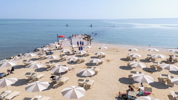 Een strand met vele parasols en ligstoelen, een rotsachtige zeebaan en een rustige zee.