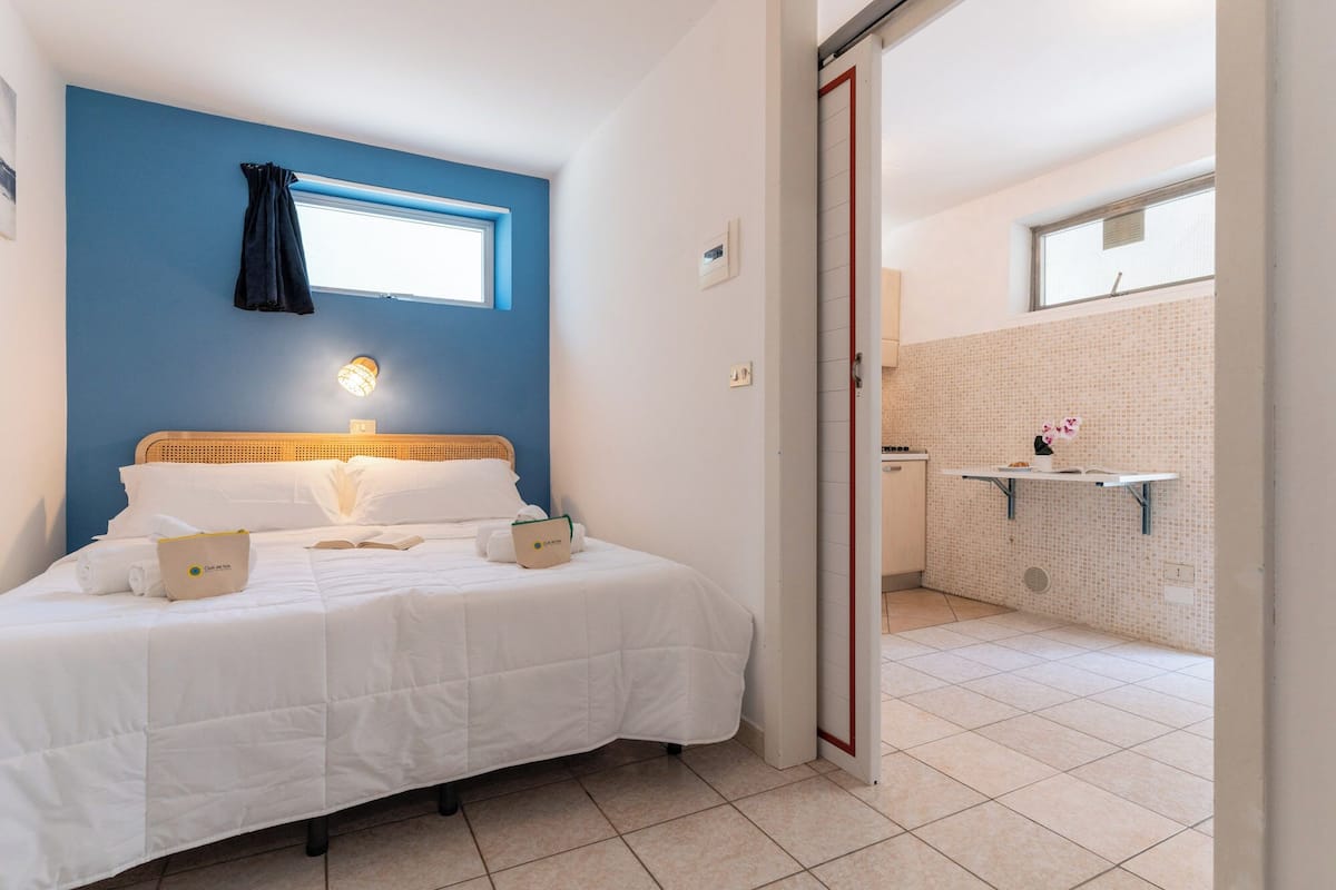 A hotel room with a bed, a bedside lamp, a window with curtains, a bathroom with a shower, and a tiled floor.