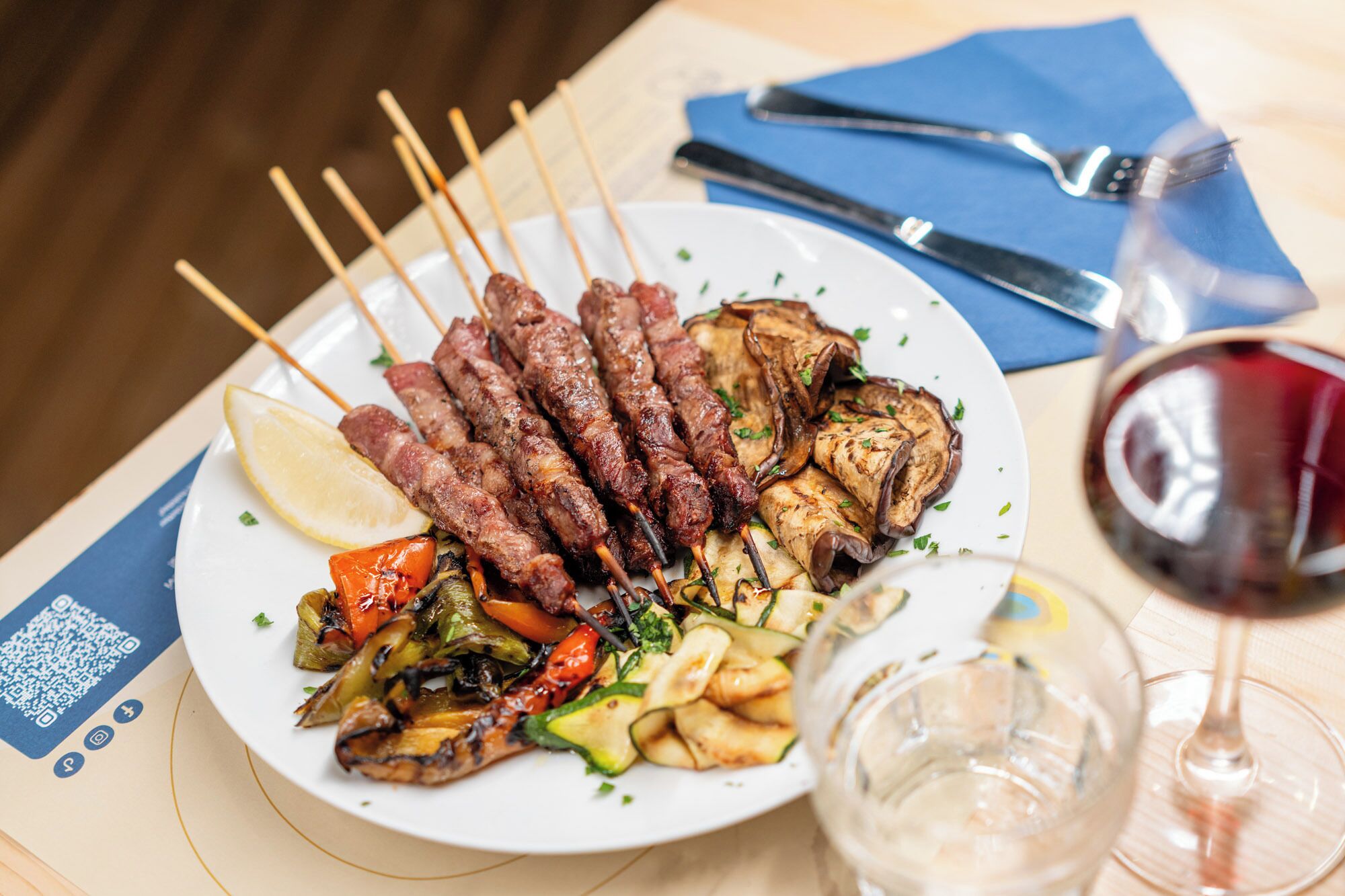 A plate of skewered meat, grilled vegetables, and a glass of red wine on a table.