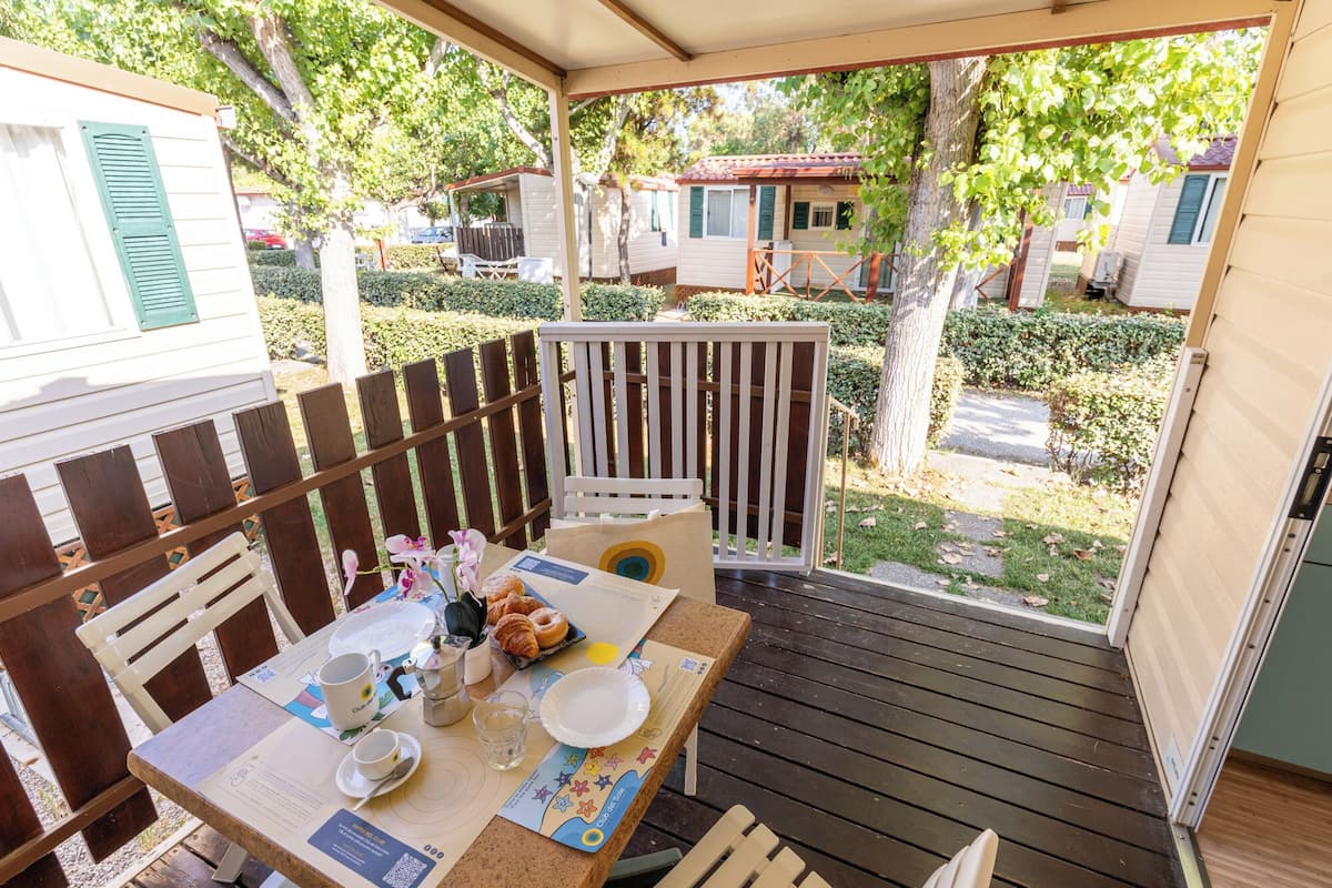 A covered outdoor seating area with a table set for breakfast, surrounded by a wooden deck and a white fence.